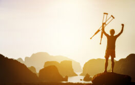 Man with crutches stands in winner pose with rised hands on mountains and islands background. Space for text